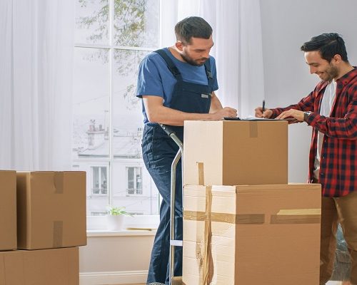 Happy New Homeowner Welcomes Professional Mover with Hand Truck full of Cardboard Boxes, Receives His Goods and Signs on Clipboard
